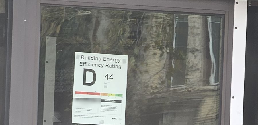 NYC Building Energy Efficiency Rating label showing grade D and score 44, posted on an old single-glazed aluminum storefront door in Brooklyn