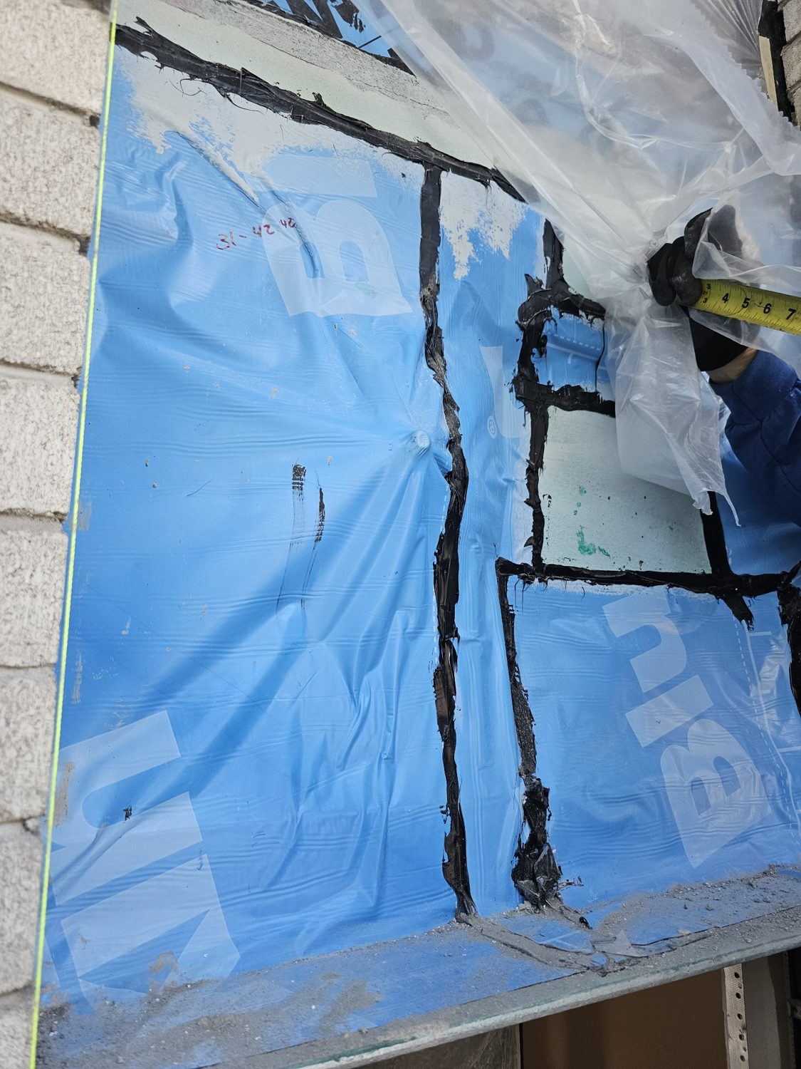 A blue self-adhered waterproofing membrane at a window opening that is wrinkled, loose from the wall and patched with black mastic