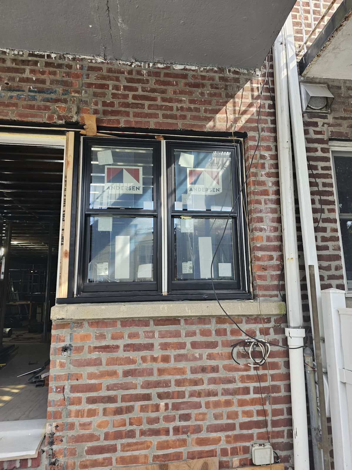 Pair of Andersen 400 Series double-hung windows with black exterior set in a brick opening during installation, factory labels and protective film still on the glass