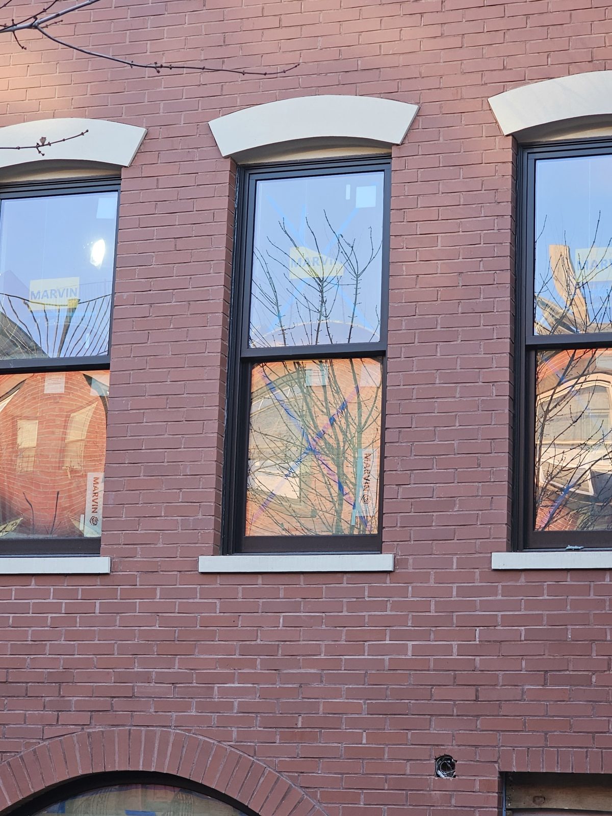 Close view of a dark one-over-one Marvin double-hung window with factory labels under a curved stone lintel in red brick at 126 Washington Avenue, Clinton Hill