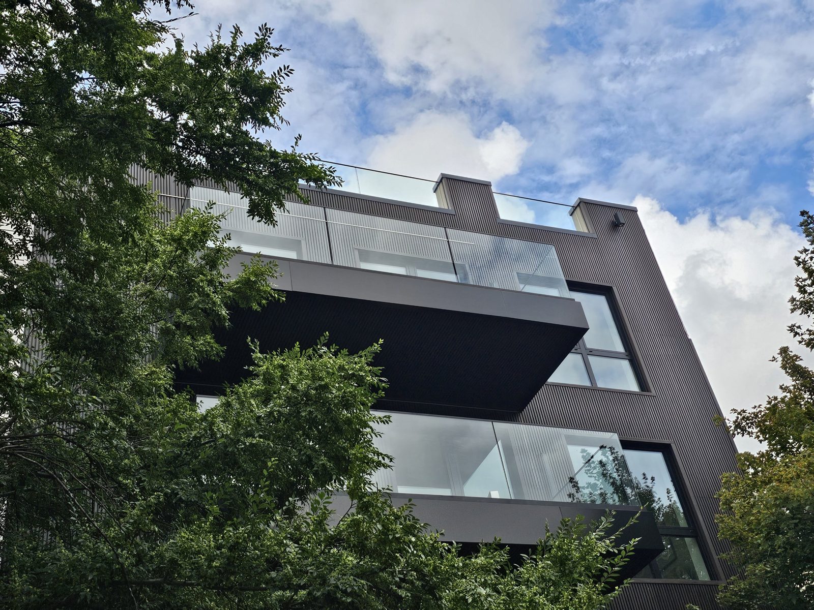 Street elevation of The Dodworth Collective at 17 Dodworth Street, Bushwick, Brooklyn: charcoal brick base with black Everest aluminium windows, cantilevered balconies wrapped in dark panels with frameless glass railings, ribbed panel cladding above and a glass roof-deck parapet