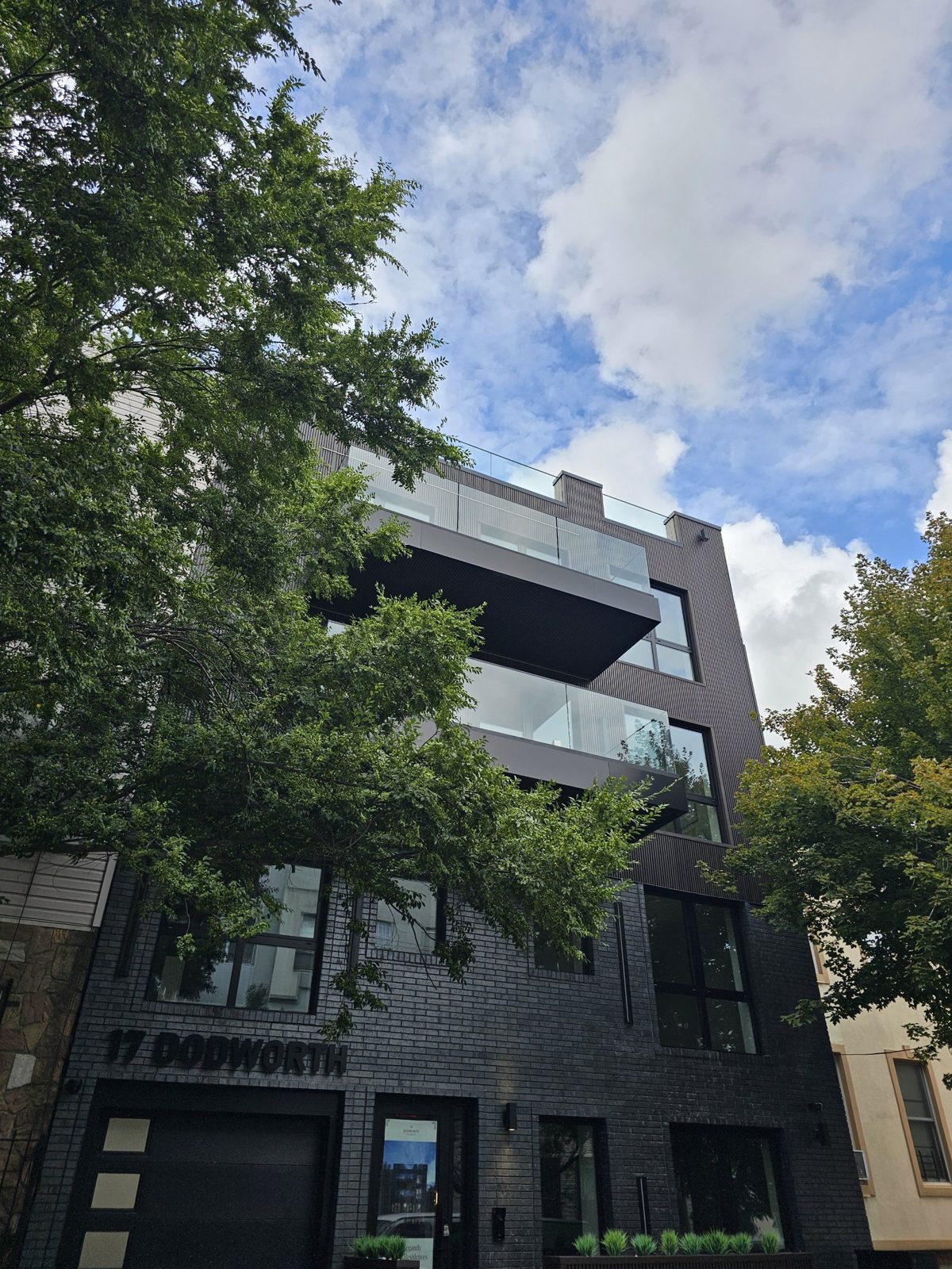 17 Dodworth Street, Bushwick, Brooklyn, seen from the sidewalk below: black aluminium windows set flush in charcoal brick, the 17 Dodworth lettering, and the panel-clad balconies with glass railings above