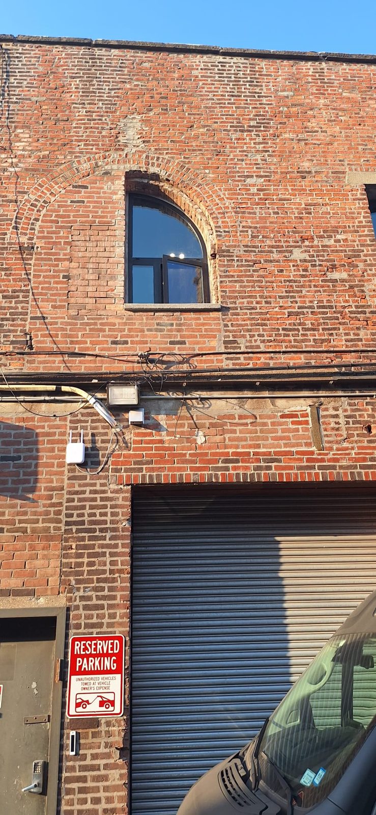Brick warehouse facade at 185 Van Dyke Street, Red Hook, with a custom half-arch aluminium window above a roll-down gate, the bricked-in half of the original arch visible beside it