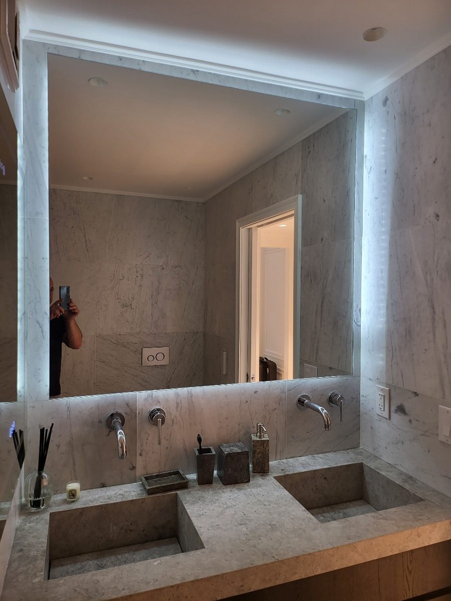 Custom LED backlit mirror over a double stone vanity with wall-mounted faucets in a Carrara marble bathroom at 95 Greene Street, SoHo