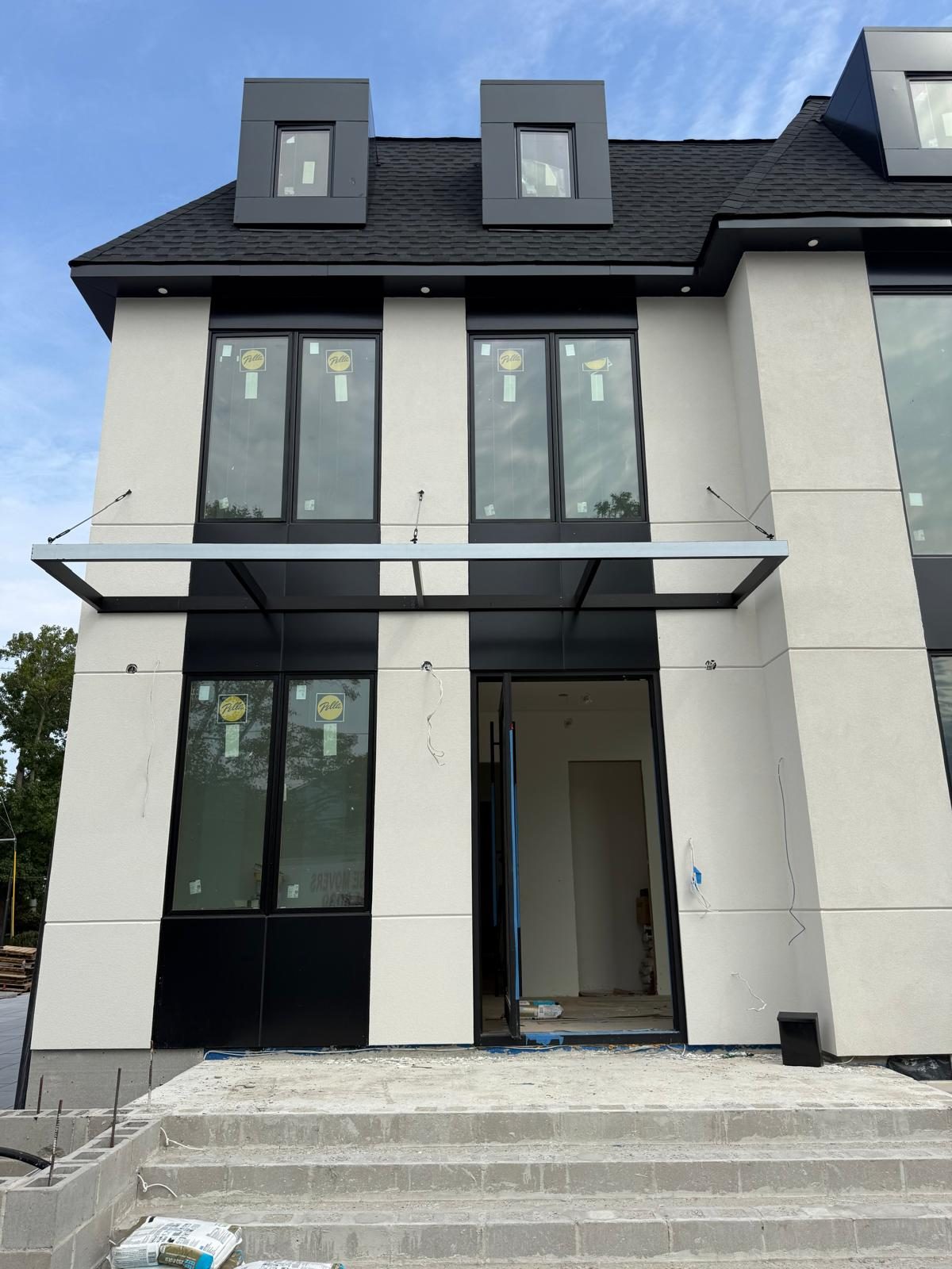Front elevation of a new custom house in Woodmere, Long Island: white stucco, black aluminum composite panels, Pella windows, a cantilevered steel entrance canopy and two box dormers