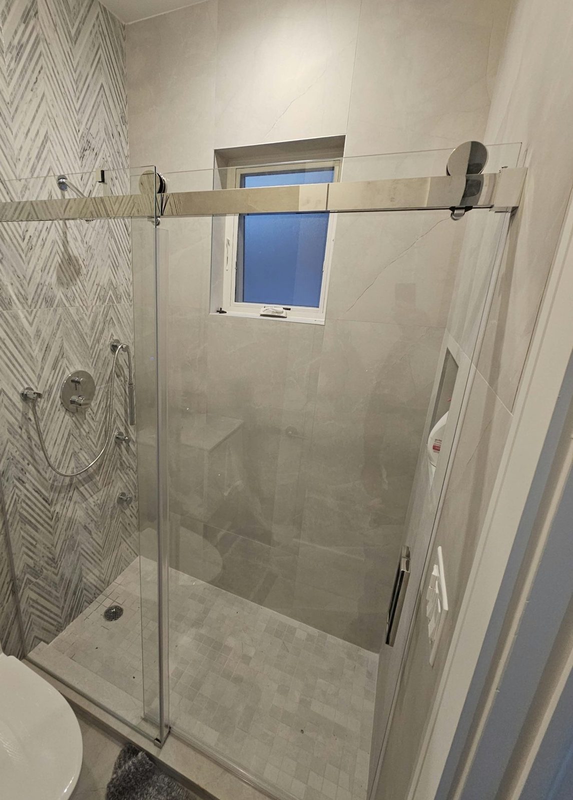 Frameless sliding glass shower door with polished bar and rollers, closed, in a tiled bathroom in Borough Park, Brooklyn