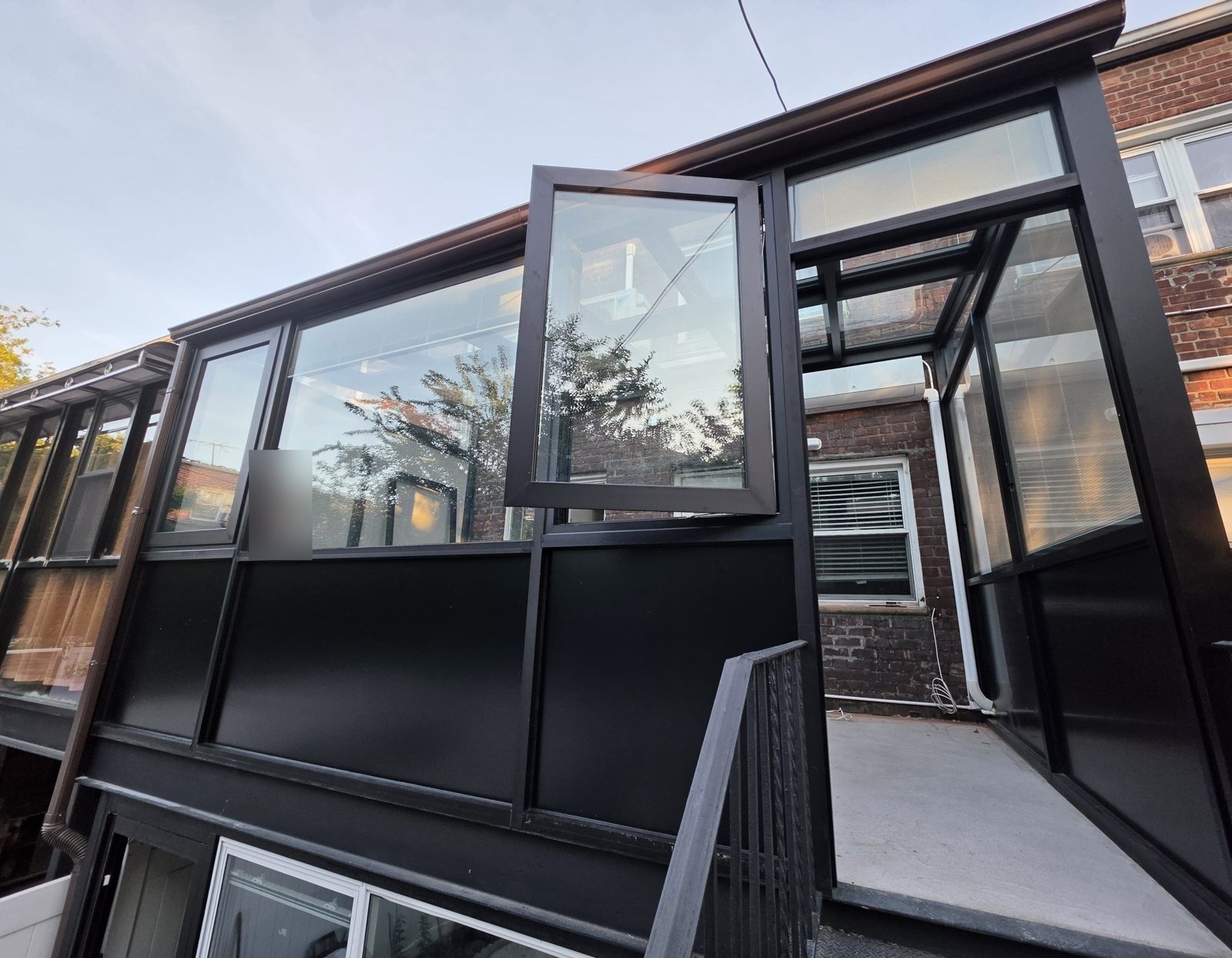 Black aluminum sunroom with glass walls, solid lower panels and an open casement window on the rear terrace of a brick house in Brooklyn, NY