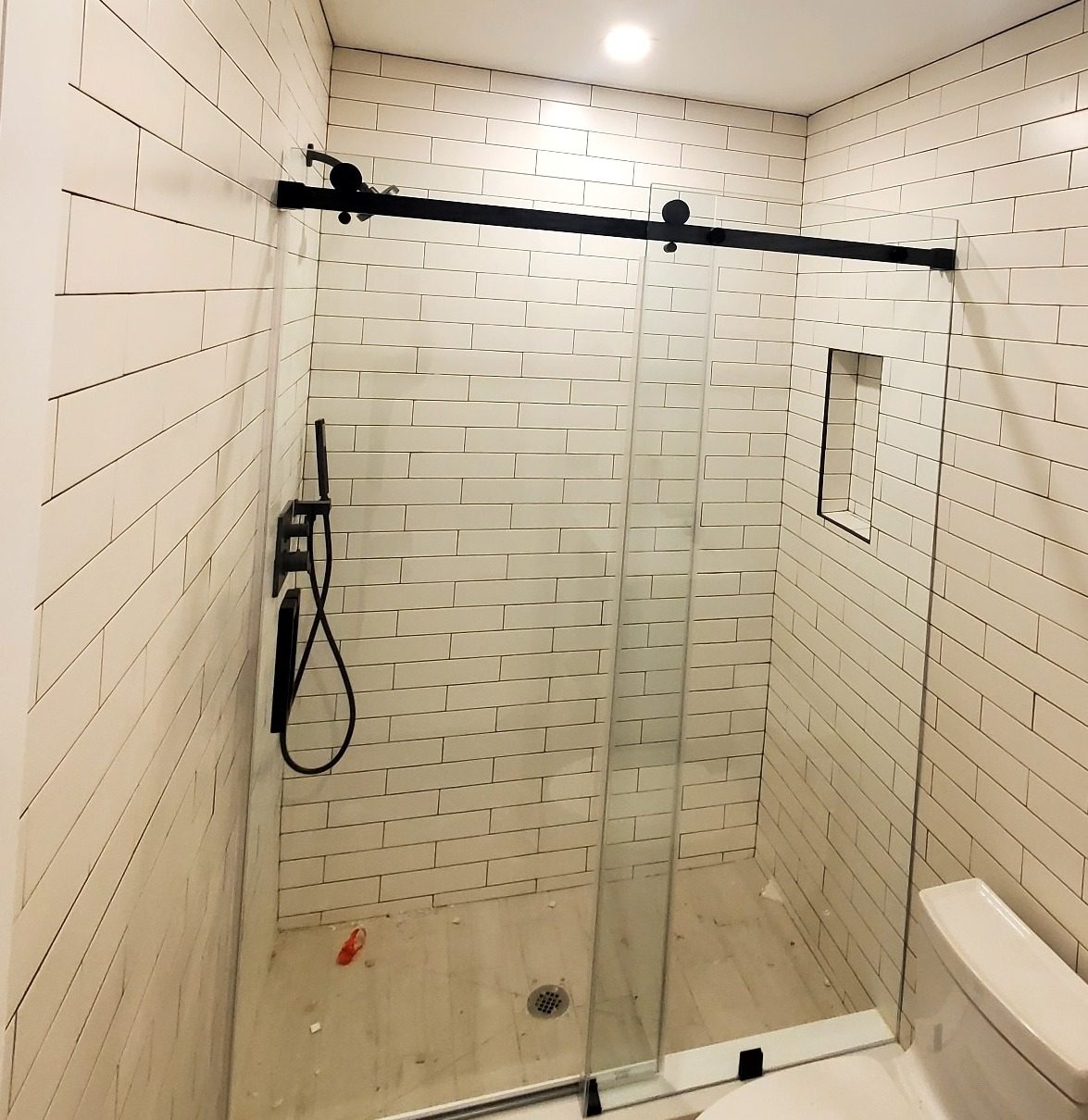 Frameless sliding glass shower door with black bar and rollers in a white subway-tile bathroom, Midwood, Brooklyn