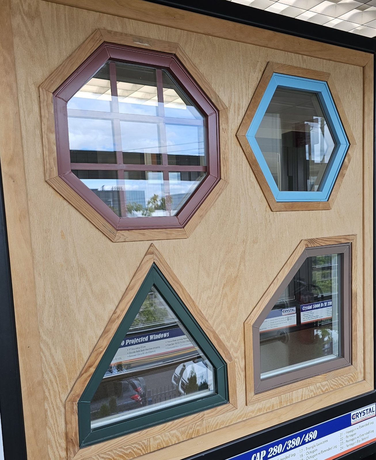 Showroom samples of aluminium shaped windows: an octagon with grids in burgundy, a hexagon in light blue, a triangle in dark green and a trapezoid in brown