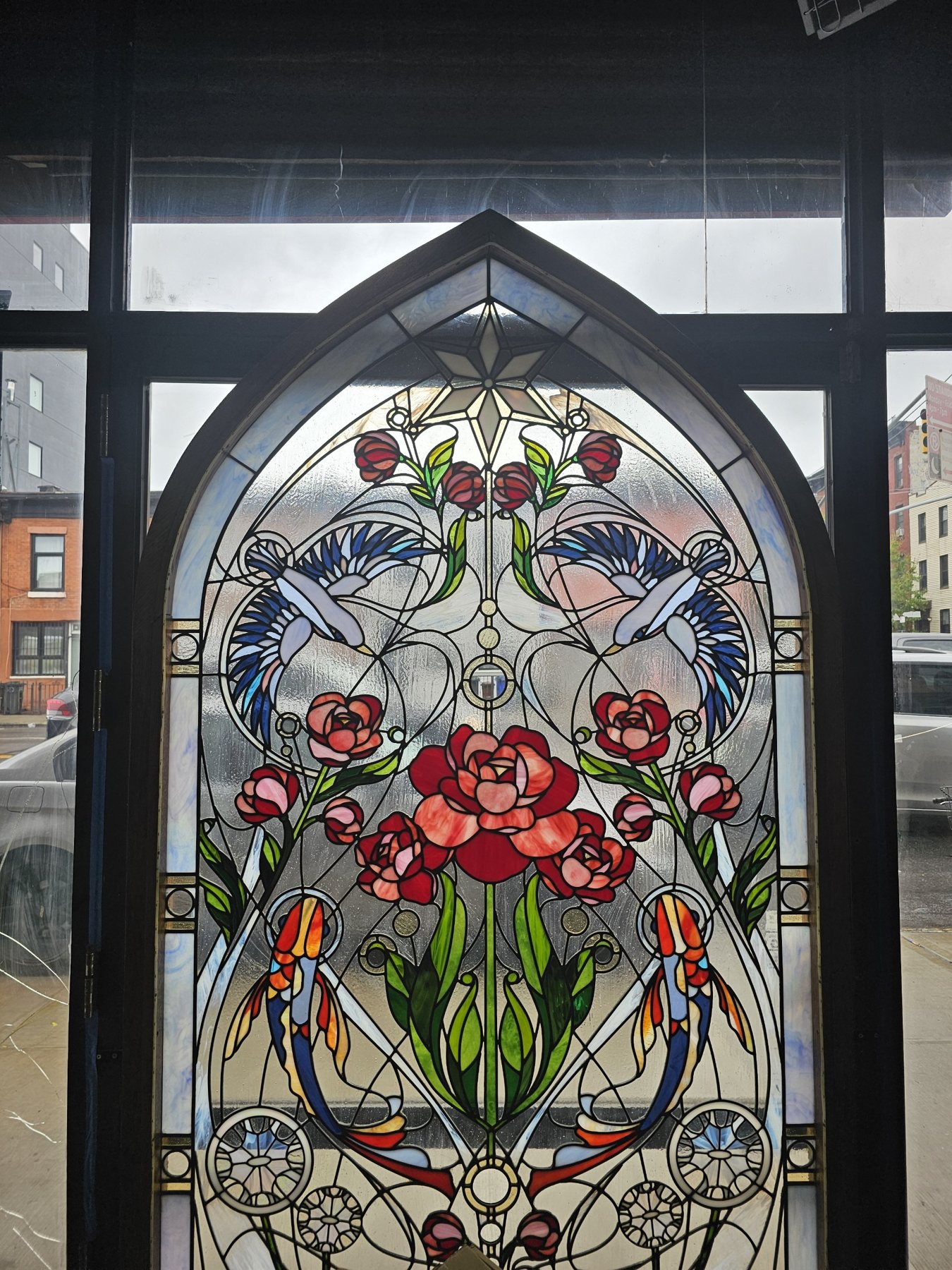 Gothic-arch stained glass panel with roses, two birds, koi fish and Art Nouveau tracery, set in a wood frame against a window