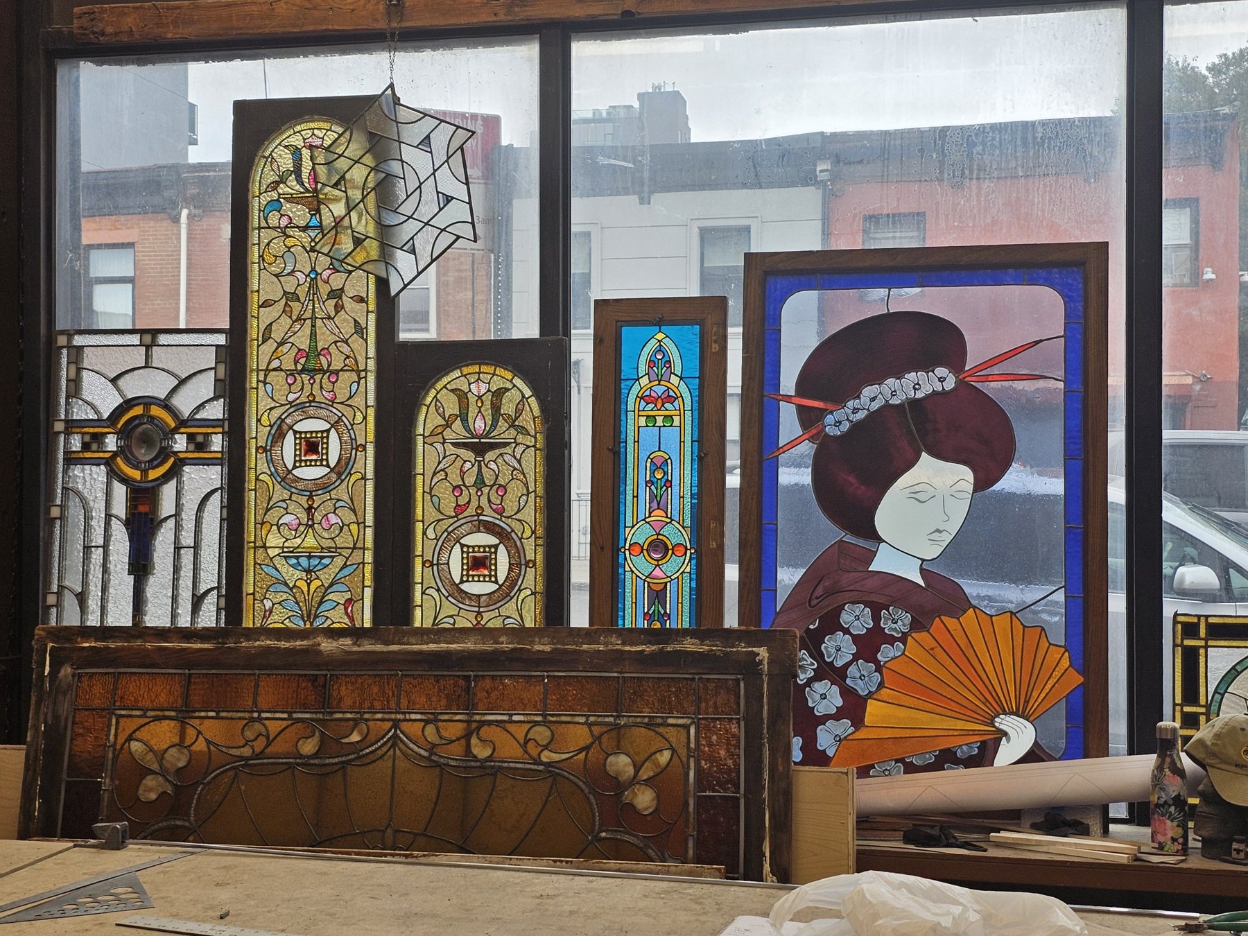 Stained glass panels standing in a studio window: Victorian arched transoms with jewel centres, a Prairie-style panel, a blue Gothic lancet and an Art Nouveau geisha panel
